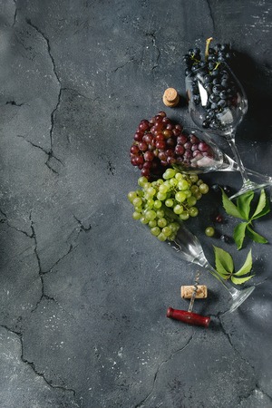 Variety of three type fresh ripe grapes dark blue, red and green in different laying wine glasses with old corkscrew and green leaves over black texture background. Top view with space.の写真素材