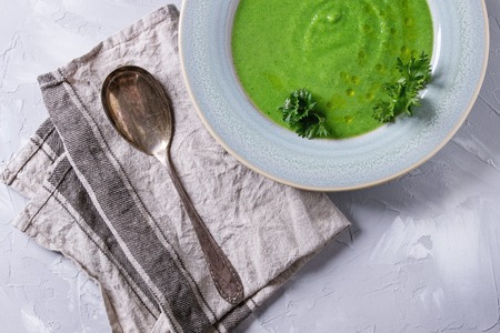 Vegetarian vegan broccoli cream soup served in blue plate with fresh parsley, broccoli, spoon, textile napkin over gray concrete background. Top view with copy space. Healthy eating.の写真素材