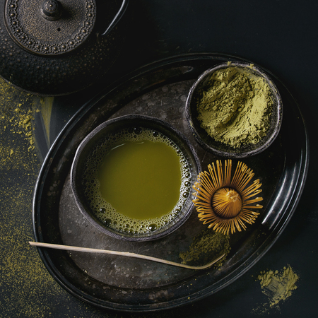 Green tea matcha powder and hot drink in black bowls standing with iron teapot, bamboo traditional tools spoon and whisk in vintage tray over dark metal background. Top view. Square imageの写真素材