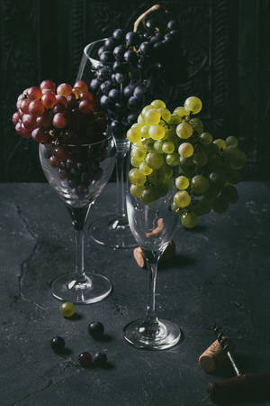 Variety of three type fresh ripe grapes dark blue, red and green in different standing wine glasses with old corkscrew over black texture background.の写真素材