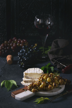 Serving board with sliced camembert cheese and baked bunch of green grapes served with glass of red wine, corkscrew, green leaves over black table. Dark rustic still lifeの写真素材