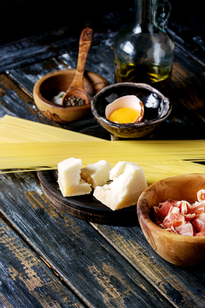 Ingredients for traditional italian pasta alla carbonara. Uncooked spaghetti, pancetta bacon, parmesan cheese, egg yolk, salt, pepper in olive wood bowls over old plank background.の写真素材