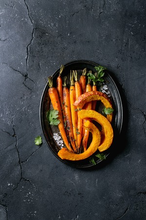 Roasted young whole carrot and sliced pumpkin with greens and sea salt. Served on vintage metal tray over black texture background. Top view with spaceの写真素材