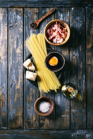 Ingredients for traditional italian pasta alla carbonara. Uncooked spaghetti, pancetta bacon, parmesan cheese, egg yolk, salt, pepper in olive wood bowls over old plank background. Top view with spaceの写真素材