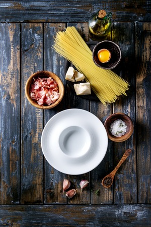 Ingredients for traditional italian pasta alla carbonara. Uncooked spaghetti, pancetta bacon, parmesan cheese, egg yolk, salt, pepper, empty plate over old plank background. Top view with spaceの写真素材