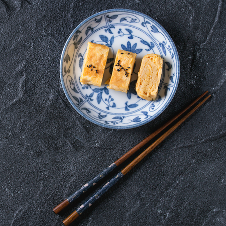 Japanese Rolled Omelette Tamagoyaki sliced with black seasame seeds and soy sauce, served in blue white ornate ceramic plate with chopsticks over black stone background. Top view. Square imageの写真素材