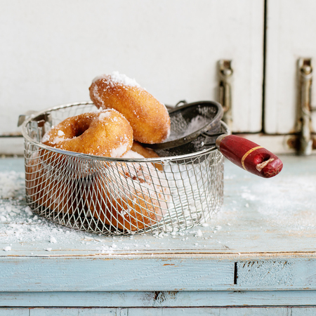 Homemade donuts with sugar powder in frying basket served with vintage sieve on blue wooden table. Rustic style, day light. Square imageの写真素材