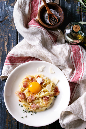 Italian traditional pasta alla carbonara with yolk, pancetta bacon, parmesan cheese, thyme, served in white plate on textile over old wooden plank background. Ingredients above. Top view with spaceの写真素材