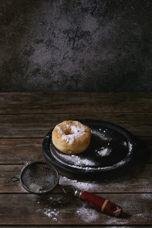 Homemade donut with sugar powder on black serving board with vintage sieve on old wooden plank table. Dark rustic style.の写真素材