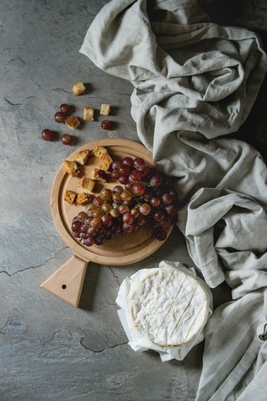 Bunch of red grapes, camembert cheese, croutons served on wooden serving board over gray kitchen table with textile linen. Rustic style. Top view with spaceの写真素材