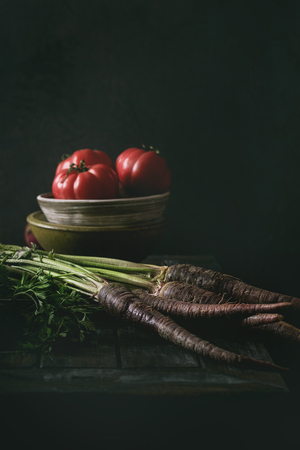 Bowl of tomatoes with purple carrot, onion on old wooden kitchen table. Dark rustic still life.の写真素材