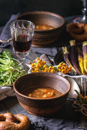 Fall holiday table decoration setting with bowls of hot carrot potato soup, baking pumpkin, carrot, garlic, fresh coriander, pretzels bread, red wine, berries, candle over wooden table. Rustic styleの写真素材