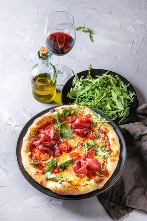 Whole homemade pizza with cheese and bresaola, served on black plate with fresh arugula, olive oil, glass of red wine and textile napkin over gray concrete texture background.の写真素材