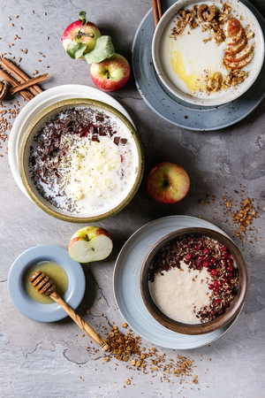 Variety bowls of milk cereal porridge with different additives, served with apples, berries and seeds over gray kitchen table. Top view with spaceの写真素材