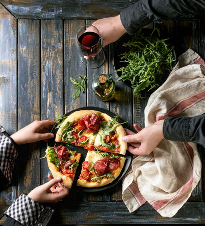 Hands taking sliced homemade pizza with cheese and bresaola, served on black plate with fresh arugula, olive oil, glass of red wine and kitchen towel over old wooden plank background. Flat lay.の写真素材
