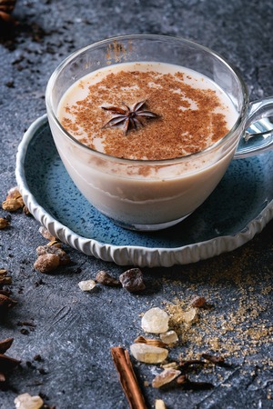 Glass cup of traditional indian masala chai tea on blue saucer with ingredients above. Cinnamon, cardamom, anise, sugar, black tea over dark texture background. Close up with space