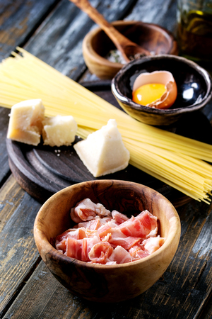 Ingredients for traditional italian pasta alla carbonara. Uncooked spaghetti, pancetta bacon, parmesan cheese, egg yolk, salt, pepper in olive wood bowls over old plank background.の写真素材