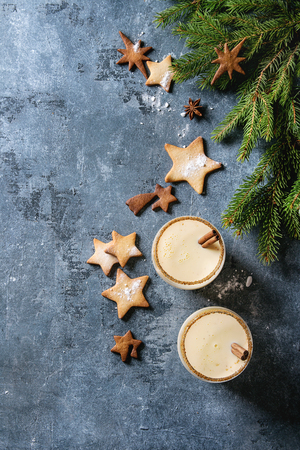 Eggnog Christmas milk cocktail with cinnamon, served in two glasses with shortbread star shape sugar cookies different size, fir branch over blue texture background. Top view with spaceの写真素材