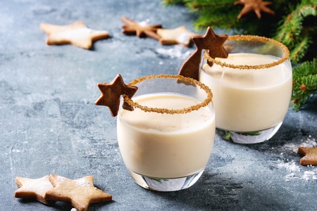 Eggnog Christmas milk cocktail with cinnamon, served in two glasses with shortbread star shape sugar cookies different size, decor toys, fir branch over blue texture background.の写真素材