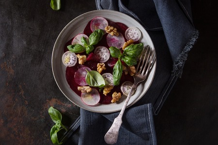 Vegan beetroot carpaccio salad with radish, basil, olive oil and walnuts in ceramic plate over dark rusty metal background. Top view, space. Dark rustic style, healthy eatingの写真素材