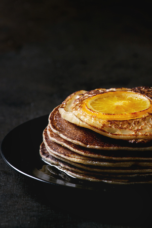 Stack of homemade pancakes with fried orange, served with mapple syrup on black plate over dark background. Close upの写真素材