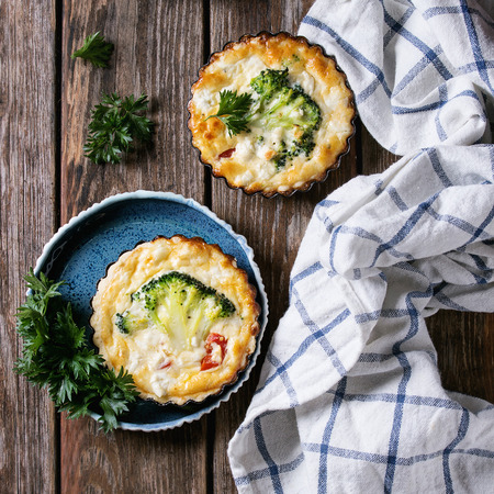 Baked homemade quiche pie in mini metal forms served with fresh greens with plate and kitchen towel on old plank wooden background. Flat lay with space. Ready to eat. Square imageの写真素材