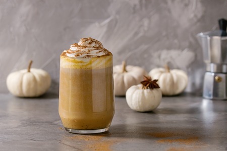 Glass of spicy pumpkin latte with whipped cream and cinnamon standing on gray kitchen table with coffeepot and decorative white pumpkins. Coffee beans and spices above. Atmospheric breakfastの写真素材