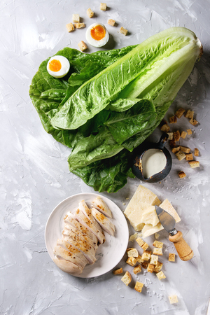Ingredients for cooking classic Caesar salad. Sliced baked chicken breast, green roman salad, parmesan cheese, boiled egg, croutons, salt, jug of sauce over gray texture background. Top view, space.の写真素材