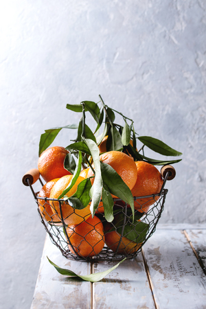 Ripe organic clementines or tangerines with leaves in basket standing on white wooden plank table with gray wall as background. Rustic style. Healthy eatingの写真素材