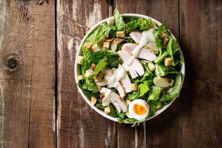 Classic Caesar salad with grilled chicken breast and half of egg in white ceramic plate. Served with croutons and dressing over old wooden background. Top view, space.の写真素材