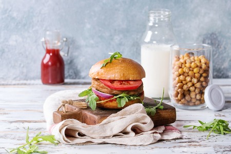 Homemade burger in classic bun with tomato sauce, arugula, meat, cheese, onion on wood serving board with bottle of ketchup, milk, roast chickpeas over white wooden plank table.の写真素材