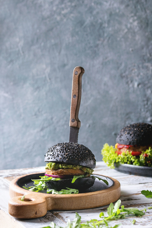 Homemade burger in black and white buns with avocado, tomato sauce, lettuce, arugula, cheese, onion on wood serving board over white wooden plank table. Rustic style. Homemade fast food.の写真素材