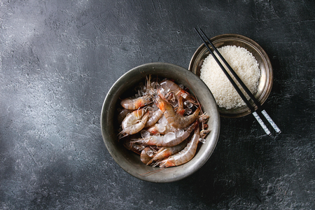 Raw whole fresh uncooked prawns shrimps on ice in vintage metal bowl with plate of raw rice and chopsticks over black texture background. Top view with copy spaceの写真素材