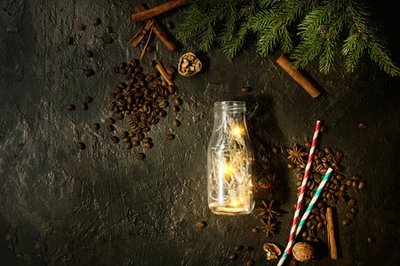Christmas lights garland in glass bottle with coffee beans, spices, nuts, cocktail tube, fir tree branches in wooden tray over dark texture background. Christmas holiday mood card. Top view, spaceの写真素材