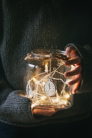 Boy in gray wool pullover holding in arms Christmas lights garland and vintage clock at 12 in glass jar. Christmas holiday mood.の写真素材