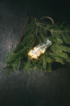 Christmas lights garland in glass bottle on green fir tree branches in wooden tray over dark texture background. Christmas holiday mood card. Top view with spaceの写真素材