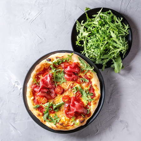 Whole homemade pizza with cheese, served on black plate with fresh arugula over gray concrete texture backgroundの写真素材