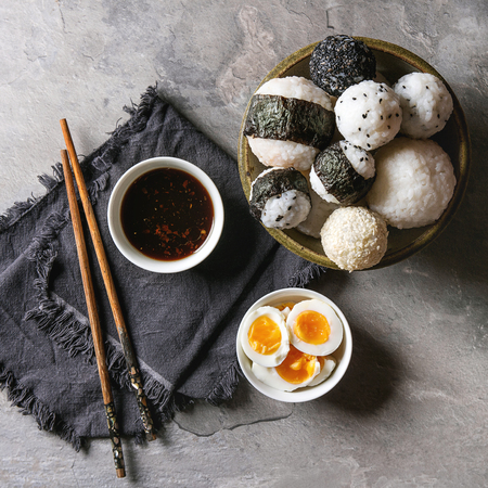Bowl with different size rice balls with black sesame and seaweed nori, served with soft boiled eggs, soy sauce, chopsticks over gray table. Asian style dinner. Top view with space. Square imageの写真素材
