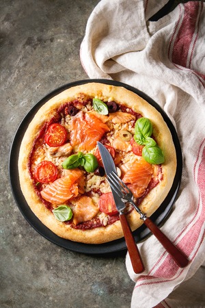 Traditional pizza with smoked salmon, cheese, tomatoes and basil served on black plate with kitchen towel over old dark metal background. Top view with space. Rustic styleの写真素材