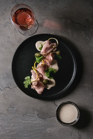 Vitello tonnato italian dish. Thin sliced veal with tuna sauce, capers and coriander served on black plate with glass of rose wine over gray texture kitchen table. Top view, spaceの写真素材
