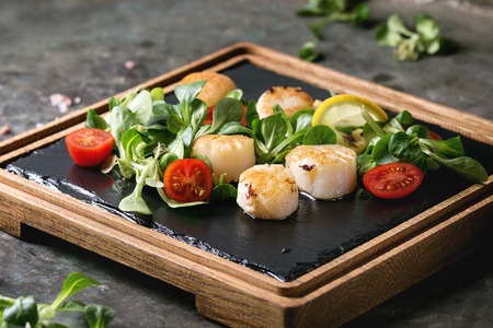 Fried scallops with lemon, cherry tomatoes and green salad served on wooden black slate serving board over old dark metal background. Close upの写真素材