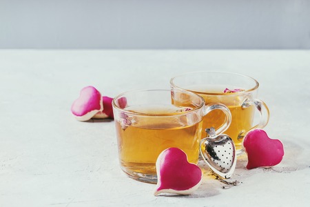 Love Valentines day greeting card with two glasses of hot tea and heart shape homemade cookies with pink icing and tea strainer over white table. Copy spaceの写真素材