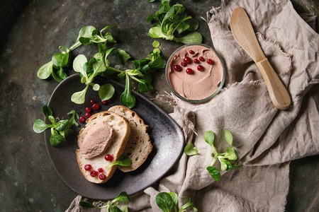 Chicken homemade liver paste or pate in glass jar with sliced whole grain bread, wood knife, cranberries, green salad served on ceramic plate with textile over dark metal background. Top view, spaceの写真素材