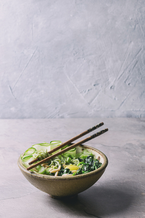Ceramic bowl with vegetarian green pea noodles with sliced cucumber, celery, spinach, quail egg yolk, pine nuts served with wooden chopsticks. Gray kitchen table. Copy space. Healthy eating. Tonedの写真素材