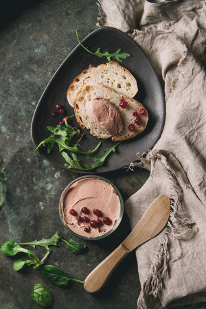 Chicken homemade liver paste or pate in glass jar with sliced whole grain bread, wood knife, cranberries, green salad served on ceramic plate with textile over dark metal background. Top view, spaceの写真素材