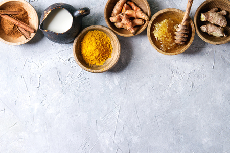 Ingredients for turmeric latte. Ground turmeric, curcuma root, cinnamon, ginger, honeycombs in wooden bowls, jug of milk over grey texture background. Top view, copy spaceの写真素材