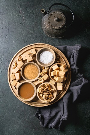 Salted caramel fudge candy served on wooden board with fleur de sel, caramel sauce, brown cane sugar and caramelized walnuts in wood bowls, iron teapot over black texture background. Top view, space.の写真素材