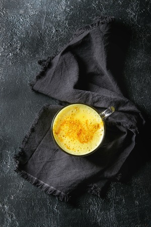 Cup of ayurvedic drink golden milk turmeric latte with curcuma powder on dark textile napkin over black texture background. Top view, spaceの写真素材