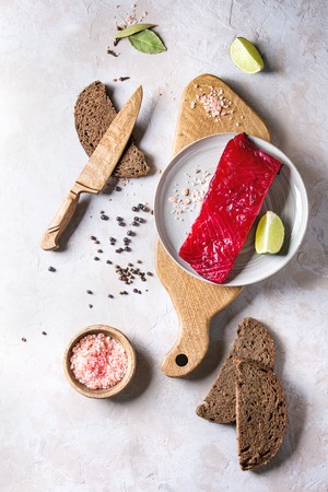 Piece of beetroot marinated salted salmon with sliced rye bread, pink salt, pepper and lime served on wooden cutting board over grey texture background. Top view, spaceの写真素材