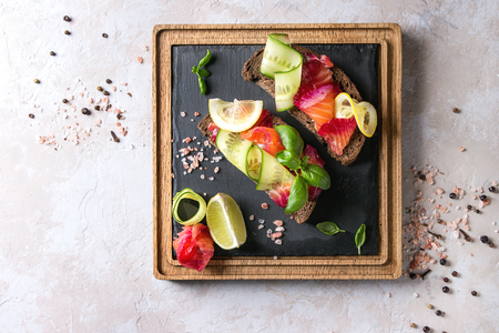 Sliced beetroot marinated salmon sandwiches with rye bread, cucumber, basil and lemon served on wooden slate cutting board over grey texture background. Top view, spaceの写真素材